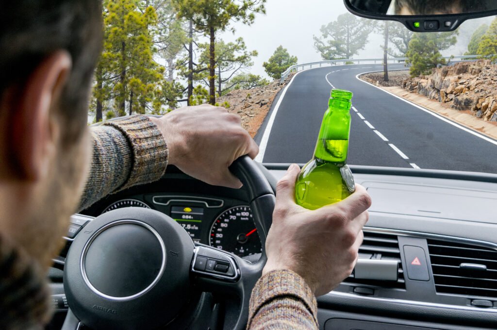 Drunk Young Man Driving A Car On The Road With A Bottle Of Beer.